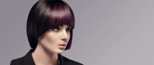 A person with a short, dark bob haircut featuring purple highlights exemplifies modern hair styles, wearing a black outfit and looking off to the side against a gray background.