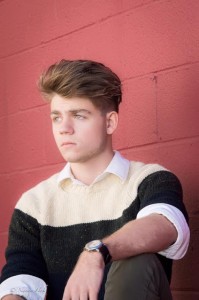 A young person with short, stylish hair and a striped sweater sits against a red brick wall, contemplating hair styles as they look thoughtfully into the distance.