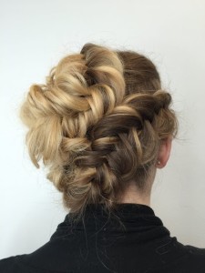 A rear view of a woman with blonde hair styled in an elaborate updo featuring intertwined braids and curls, showcasing one of the most intricate hair styles. She is wearing a black top.