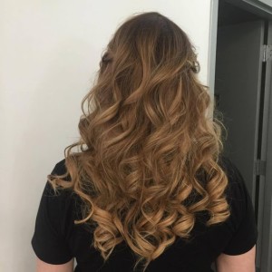 A person with long, wavy, light brown hair seen from the back, wearing a black shirt and showcasing one of the latest hair styles, stands indoors near a grey door.
