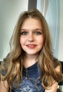 A young person with long, wavy, light brown hair showcasing one of the latest hair styles and blue eyes smiles at the camera, wearing a dark blue shirt. The background features a light-colored wall with abstract patterns.