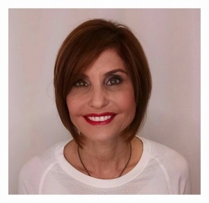 A woman with short brown hair styles, wearing a white top, is smiling at the camera against a plain background.