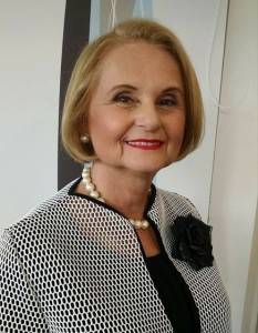An older woman with short blonde hair styled elegantly, a pearl necklace, and a white and black patterned jacket with a black flower brooch, smiles at the camera.