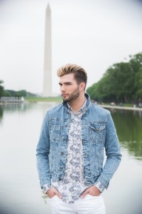 A man with blond hair and a trimmed beard stands with hands in pockets in front of a reflecting pool with the Washington Monument in the background. Showcasing one of the latest hair styles, he is wearing a denim jacket over a floral shirt.