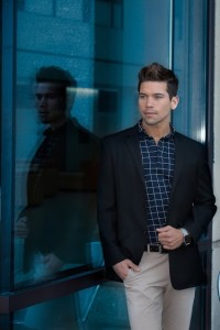 A man in a black blazer and checkered shirt stands next to a glass window, his reflection visible. With his hand in his pocket, he looks away from the camera, showcasing one of today's sleek hair styles.