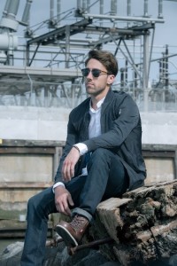 A man wearing sunglasses and casual clothing sits on rocks in an industrial area, showcasing various hair styles with steel structures and machinery visible in the background.