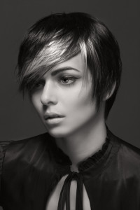 Black and white portrait of a person with short, dark hair featuring light streaks, wearing a high-collared, buttoned shirt. Their gaze is off-frame with a neutral expression, showcasing timeless hair styles.