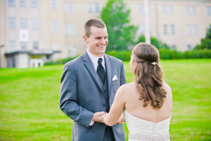 A groom in a gray suit and a bride in a white dress hold hands and smile at each other outdoors, with their elegant hair styles perfectly complementing the scenic backdrop of a building and green lawn.