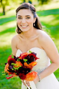 A smiling bride in a white strapless wedding dress, showcasing one of the latest hairstyles, holds a bouquet of orange, red, and yellow flowers while standing outdoors.