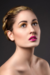 A woman with gold-accented hair and makeup, showcasing one of the latest hair styles, gazes upward against a dark background. She wears bold pink lipstick and has a neutral expression.