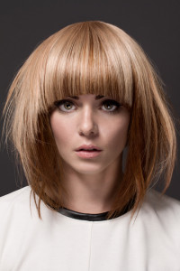 A person with shoulder-length, straight, light brown hair styled with blunt bangs showcases one of the chicest hair styles, wearing a white top against a dark background.
