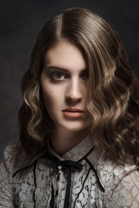 Woman with wavy brown hair styled elegantly, and a serious expression, wearing a white lace blouse with a black ribbon tie, posed against a dark background.