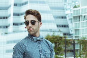 A man sporting stylish hair, sunglasses, a denim shirt, and a bow tie stands outdoors in an urban area with modern glass buildings in the background.