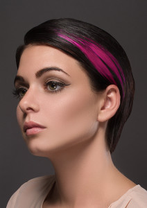 Close-up of a person with short dark hair styles featuring a prominent pink streak, light makeup, and a neutral expression, set against a dark background.