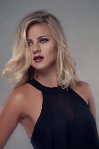 A person with shoulder-length blonde hair styles and red lipstick is wearing a black sleeveless top, looking slightly to the side against a neutral background.