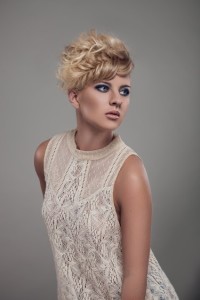 A person with short, curly blonde hair and blue eye makeup is showcasing one of today's trendy hair styles while wearing a sleeveless, lacy white top. They are posing against a grey background, looking to the side.