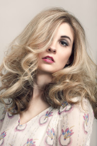 A person with long, wavy blonde hair partially covering one eye, showcasing one of the trendiest hair styles, wears a light-colored shirt with a colorful pattern while posing against a plain background.