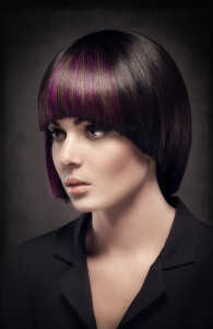 A woman with a short, straight bob haircut featuring purple highlights, showcasing one of today's trendy hair styles, is wearing a black shirt and facing left in a dimly lit setting.