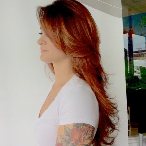 A woman with long, wavy, light brown hair wearing a white t-shirt stands in profile. She has a tattoo with floral designs on her right arm.