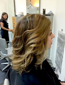 A woman with wavy, shoulder-length hair sits in a salon chair, facing right. Another person is seen in the background of the salon.