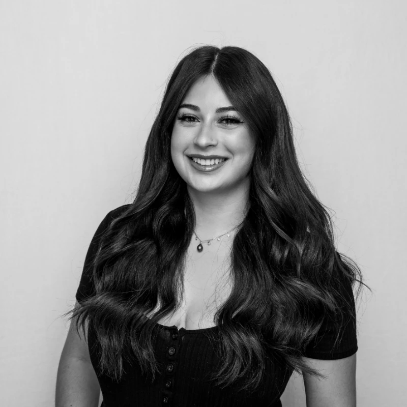 A woman with long, wavy hair is smiling at the camera. She is wearing a black top and a necklace. The image is in black and white.