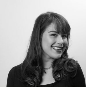 Black and white portrait of a smiling person with long, wavy hair and bangs, wearing a dark top and a thin necklace. They are looking slightly to the right.