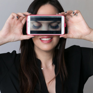 A woman holds a smartphone horizontally in front of her eyes, displaying an image of closed eyes with long eyelashes. She is smiling and wearing a black top.