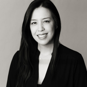 Black and white portrait of a woman with long dark hair, smiling, wearing a dark top and a small necklace, against a plain background.