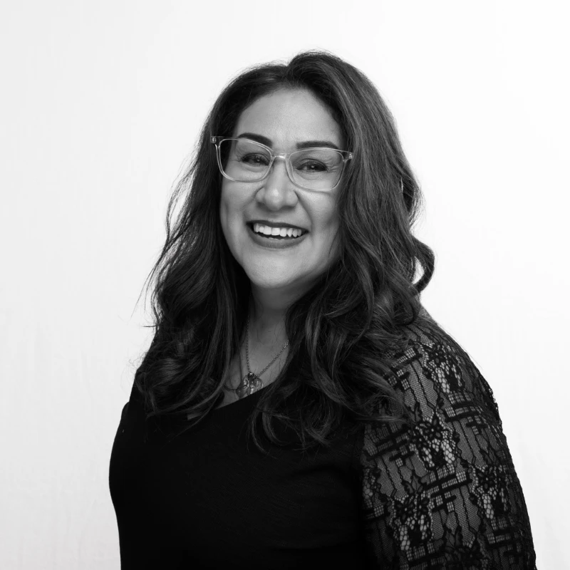 A woman wearing glasses and a lace-sleeved top smiles at the camera in a black-and-white portrait.
