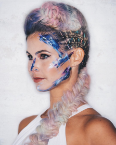 A woman with a pastel-colored braided hairstyle, created using Oribe products, and abstract blue face paint, wearing a white top, poses against a white backdrop.