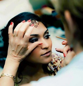 A woman at the salon is having makeup applied to her eye by another person, with her hand steadying her eyelid. She has dark hair and red nail polish, capturing a moment of beauty in today's fast-paced world.