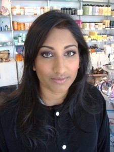 A woman stands in a store with shelves of various products in the background, reminiscent of a high-end salon today. She has long dark hair and is wearing a black top.