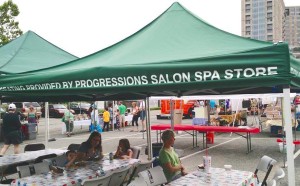 Green canopy with "PROVIDED BY PROGRESSIONS SALON SPA STORE" text covering a booth at an outdoor event with people seated at tables and others walking around. Buildings visible in the background, celebrating their recognition as one of Salon Today 200's finest.