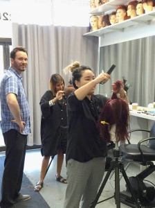 Two stylists practice on mannequins in a bustling salon today, as a person in a blue shirt observes. Shelves with additional mannequin heads are visible above.