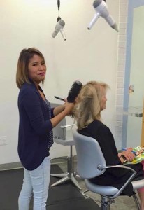 A hairdresser with a brush in hand is styling a seated woman's hair in Salon Today, where three hairdryers hang from above.