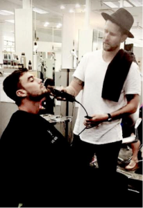 A man wearing a white t-shirt and a hat trims another man's beard in a modern salon, embodying the style and precision showcased in Salon Today's 200 top barbershops.