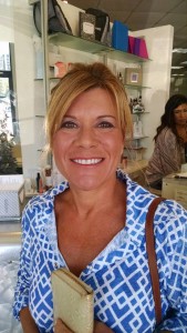 A woman with blonde hair smiles at the camera. She is wearing a blue and white patterned shirt and holding a beige clutch. A woman in the background, likely waiting for her turn, stands near shelves, making it feel like a scene straight out of Salon Today's top 200 issue.