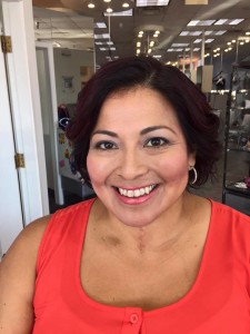 A woman with short dark hair and wearing a sleeveless red top smiles at the camera. She is standing indoors with office furniture and decor in the background, looking every bit the accomplished professional recently featured in Salon Today 200.