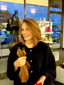 A smiling person stands indoors at a salon, holding cut sections of long brown hair while sporting a new, shorter haircut.