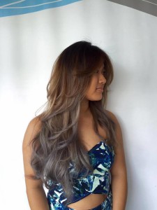 A woman with long, wavy, brown and grey ombre hair, wearing a blue patterned outfit, stands against a plain background—a look worthy of Salon Today 200.