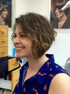 A woman with a short, wavy bob haircut smiles while sitting in the salon today. She is wearing a blue sleeveless top with a fox pattern.