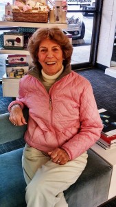 A woman with short, curly hair, dressed in a pink quilted jacket and white pants, sits smiling in a store with a basket of products and a parked car visible in the background, as if she's just stepped out from her appointment at Salon Today 200.