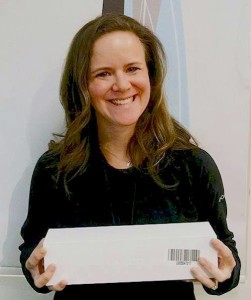 A woman with long brown hair is smiling while holding a white rectangular box with a barcode. She is wearing a dark top and standing against a plain background, looking like she just stepped out of an editorial for Salon Today 200.