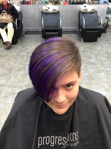 Individual with short hair featuring a purple streak, seated in a salon chair, wearing a black cape labeled "Progressions." Background shows another seated person and two vacant salon chairs, capturing the modern vibe of Salon Today 200.