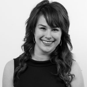A black-and-white photo of a person with long wavy hair, smiling, wearing a sleeveless top and oval-shaped earrings, against a plain background.