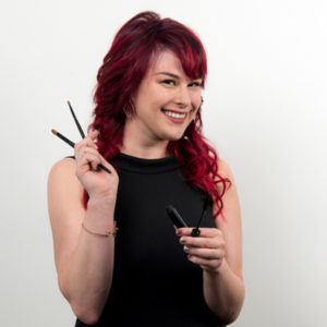 A person with red hair smiles while holding makeup brushes and a mascara wand in their hands against a plain white background.