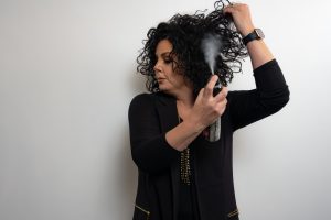 A person with curly hair is spraying a product into their hair while looking sideways.
