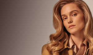 A woman with long, wavy blonde hair and light makeup looks at the camera against a gradient background. She is wearing a beige outfit.