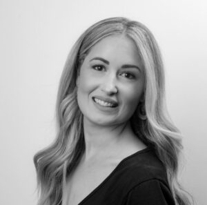 A black-and-white headshot of a woman with long, wavy hair, smiling and looking slightly to the side. She is wearing a V-neck top and earrings, exuding confidence as she pursues her career ambitions.