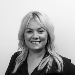 A woman with light blonde hair smiles at the camera against a plain background. She is wearing a dark top and hoop earrings, exuding confidence in her professional demeanor. The photo, captured in black and white, radiates a timeless elegance suitable for any careers portfolio.